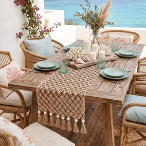 May include: A rustic wooden dining table set for an outdoor meal. The table is adorned with a macrame table runner, coral decor, and seafoam green plates. Wicker chairs with light blue cushions surround the table, with a scenic ocean view in the background.