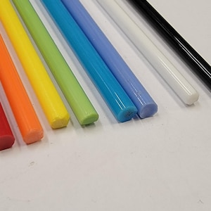 May include: A selection of brightly coloured glue sticks, including red, orange, yellow, green, blue, light blue, white, and black. The cylindrical sticks are arranged diagonally on a white surface, displaying their vibrant colours and smooth texture.