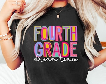 May include: Black t-shirt with the words "FOURTH GRADE dream team" in colorful, block letters. The text is outlined in various colors, including pink, yellow, blue, and purple. The shirt is a comfortable, casual style.