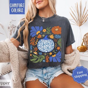Op de afbeelding: Een donkergrijs T-shirt met een blauw en wit bloemenontwerp met een pompoen met een blauw en wit patroon. De tekst "Comfort Colors" is gedrukt in de linkerbovenhoek van het shirt.
