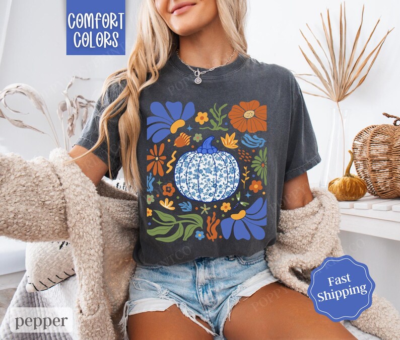 Op de afbeelding: Een donkergrijs T-shirt met een blauw en wit bloemenontwerp met een pompoen met een blauw en wit patroon. De tekst "Comfort Colors" is gedrukt in de linkerbovenhoek van het shirt.