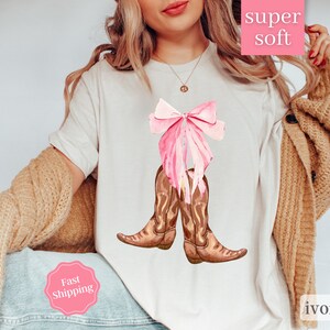 K&ouml;nnte beinhalten: Wei&szlig;es T-Shirt mit einer rosa Schleife und braunen Cowboy-Stiefeln. Der Text "super soft" befindet sich auf einem rosa Etikett und der Text "ivory" auf einem wei&szlig;en Etikett. Der Text "Fast Shipping" befindet sich auf einem rosa Etikett.