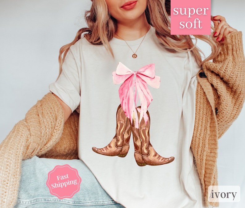 K&ouml;nnte beinhalten: Wei&szlig;es T-Shirt mit einer rosa Schleife und braunen Cowboy-Stiefeln. Der Text "super soft" befindet sich auf einem rosa Etikett und der Text "ivory" auf einem wei&szlig;en Etikett. Der Text "Fast Shipping" befindet sich auf einem rosa Etikett.