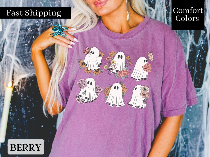 May include: Purple t-shirt with a graphic of six white ghosts holding flowers. The ghosts are surrounded by pink, orange, and yellow flowers. The shirt has the text "Fast Shipping" and "Comfort Colors" on it.