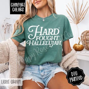 May include: Light green t-shirt with the words "Hard Fought Hallelujah" in white script. The shirt is a Comfort Colors brand and is labeled "Light Green". The image also includes a beige sweater and denim shorts.