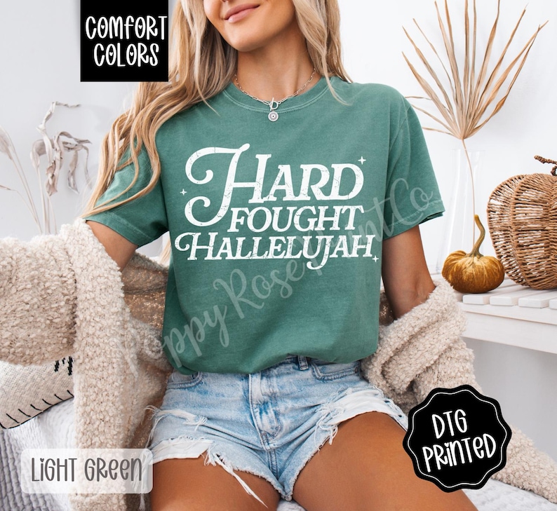 May include: Light green t-shirt with the words "Hard Fought Hallelujah" in white script. The shirt is a Comfort Colors brand and is labeled "Light Green". The image also includes a beige sweater and denim shorts.