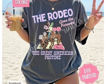以下が含まれることがあります： ピンクと白で「THE RODEO Goes On Forever」の文字が入った濃いグレーのTシャツ。カウボーイが馬に乗っている様子、さくらんぼ、ピンクのカウボーイハットのグラフィックが特徴です。「THE GREAT AMERICAN PASTIME」の文字もプリントされています。