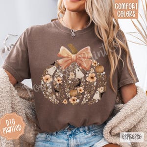 Pu&ograve; includere: T-shirt color espresso con un design a zucca con fiocco pesca, fiori, fantasmi e pipistrelli. La maglietta riporta le scritte "Comfort Colors" e "DTG Printed".
