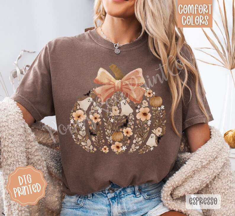 Pu&ograve; includere: T-shirt color espresso con un design a zucca con fiocco pesca, fiori, fantasmi e pipistrelli. La maglietta riporta le scritte "Comfort Colors" e "DTG Printed".