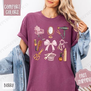 May include: A purple t-shirt with a graphic design of various hair salon tools, including a hair dryer, scissors, a hairbrush, a bowl, a spray bottle, a hair tie, a pearl necklace, a pink bow, and a salon chair.