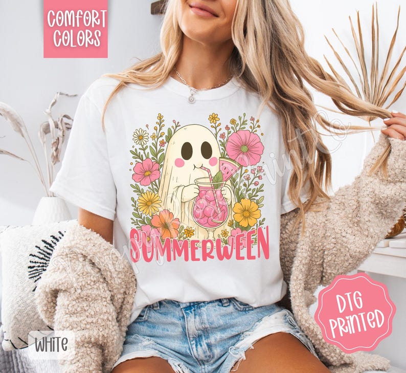May include: White t-shirt with a colorful graphic of a ghost holding a pink drink, surrounded by flowers. The word "SUMMERWEEN" is printed in pink. The shirt also has the words "COMFORT COLORS" and "WHITE" printed on it.