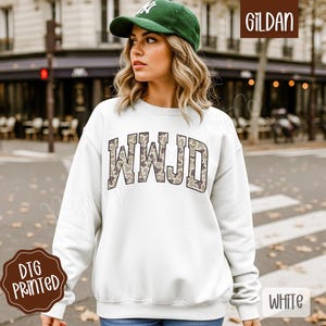 May include: White sweatshirt with the letters "WWJD" in a camouflage pattern across the chest. The sweatshirt is a Gildan brand and has a DTG printed design. The model is wearing a green cap.