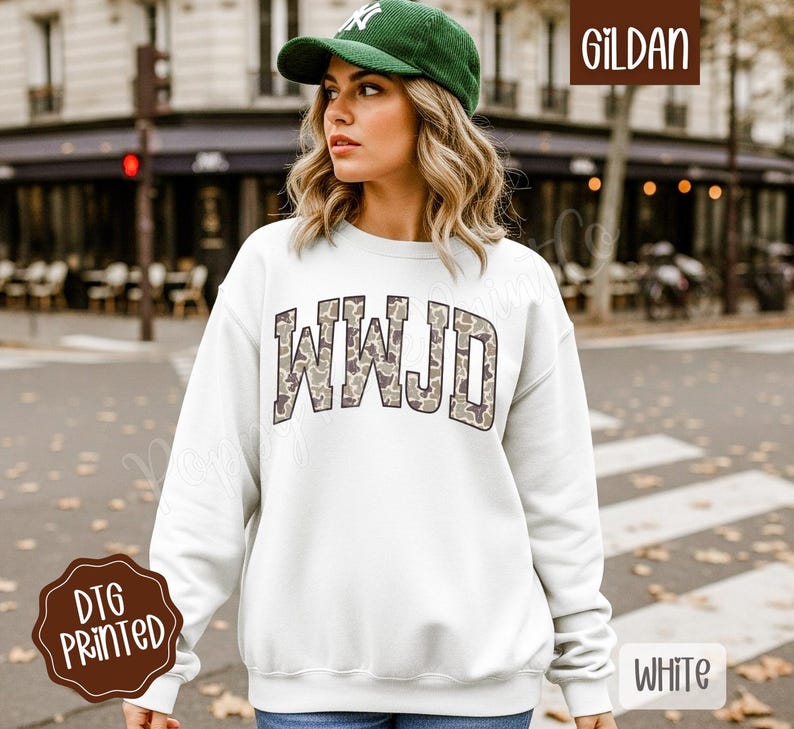 May include: White sweatshirt with the letters "WWJD" in a camouflage pattern across the chest. The sweatshirt is a Gildan brand and has a DTG printed design. The model is wearing a green cap.