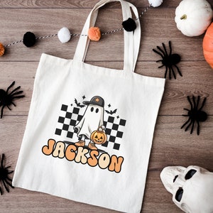 K&ouml;nnte beinhalten: Wei&szlig;e Stofftasche mit einem Cartoon-Geist, der eine Baseballkappe tr&auml;gt und eine K&uuml;rbislaterne h&auml;lt. Die Tasche ist mit dem Namen "JACKSON" in orangefarbenen und gelben Buchstaben bedruckt.