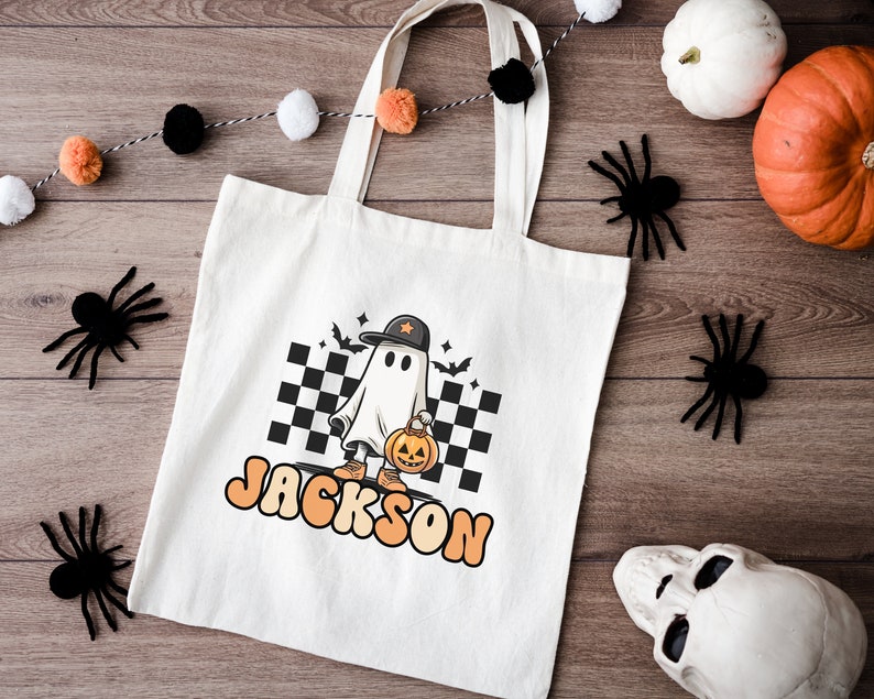 K&ouml;nnte beinhalten: Wei&szlig;e Stofftasche mit einem Cartoon-Geist, der eine Baseballkappe tr&auml;gt und eine K&uuml;rbislaterne h&auml;lt. Die Tasche ist mit dem Namen "JACKSON" in orangefarbenen und gelben Buchstaben bedruckt.