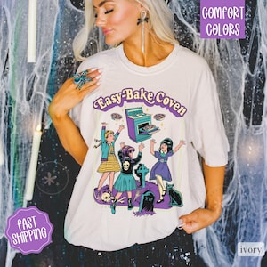 May include: An ivory t-shirt featuring a graphic print with the text "Easy-Bake Coven". The illustration depicts three children, a purple oven, a black cat, tombstones, and a skull. The design uses purple, blue, and yellow colors.
