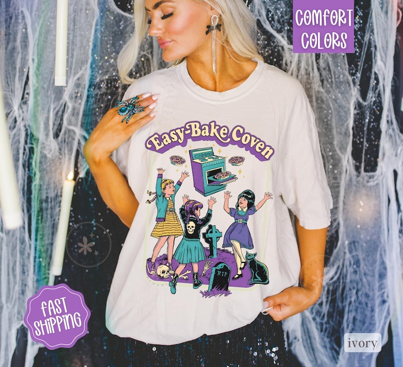 May include: An ivory t-shirt featuring a graphic print with the text "Easy-Bake Coven". The illustration depicts three children, a purple oven, a black cat, tombstones, and a skull. The design uses purple, blue, and yellow colors.