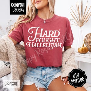 May include: Crimson-colored t-shirt with the text "Hard Fought Hallelujah" in white script. The shirt is from Comfort Colors and is DTG printed. The model is wearing denim shorts and a cream-colored cardigan.
