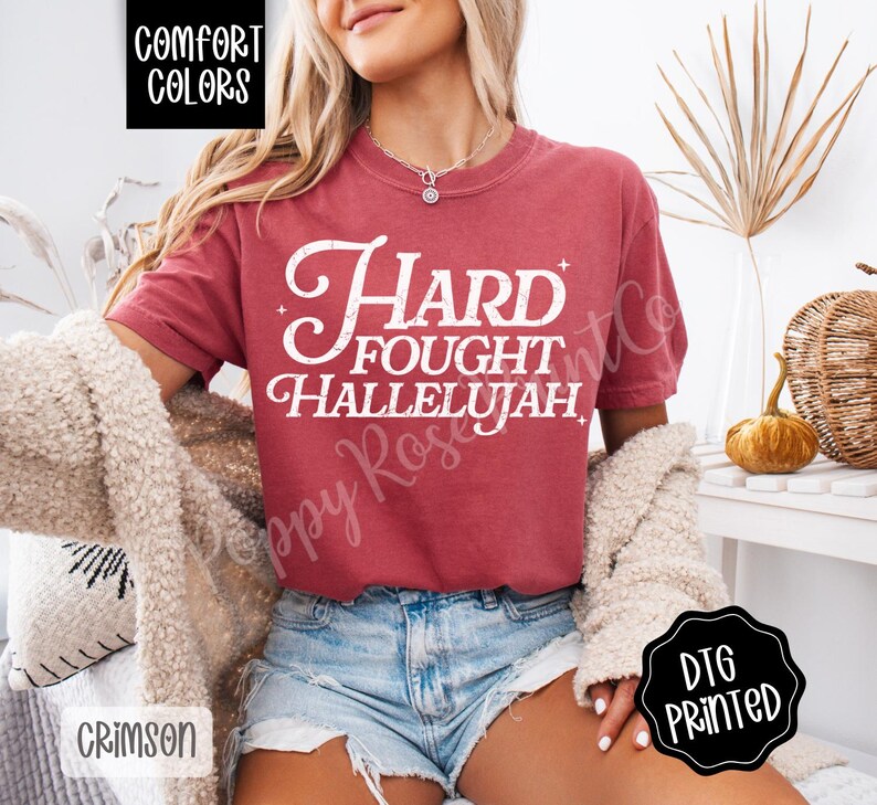 May include: Crimson-colored t-shirt with the text "Hard Fought Hallelujah" in white script. The shirt is from Comfort Colors and is DTG printed. The model is wearing denim shorts and a cream-colored cardigan.