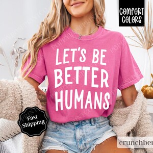 May include: A pink t-shirt with the text "Let's Be Better Humans" in white lettering. The shirt is worn by a person with long blonde hair and is styled with denim shorts.