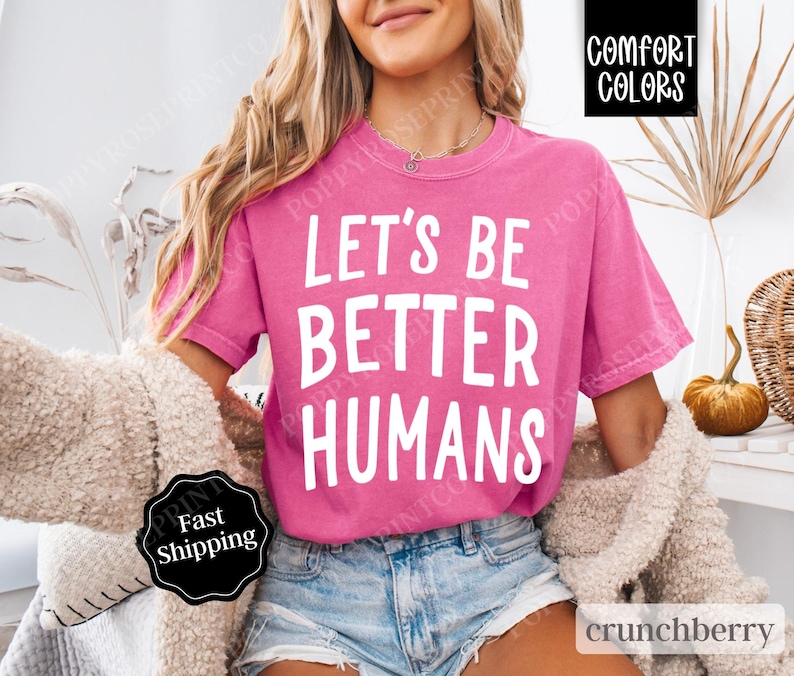May include: A pink t-shirt with the text "Let's Be Better Humans" in white lettering. The shirt is worn by a person with long blonde hair and is styled with denim shorts.