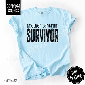 May include: Light blue t-shirt with the words "toddler tantrum SURVIVOR" in black lettering. The shirt has short sleeves and a crew neck. The words "Comfort Colors" and "Chambray" are also visible.