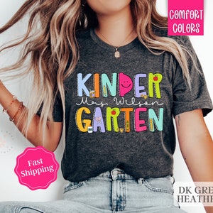 May include: A dark grey heather t-shirt with the text "KINDERGARTEN" in colorful letters with illustrations of school supplies. The text "Mrs. Wilson" is written in black below the word "KINDERGARTEN".