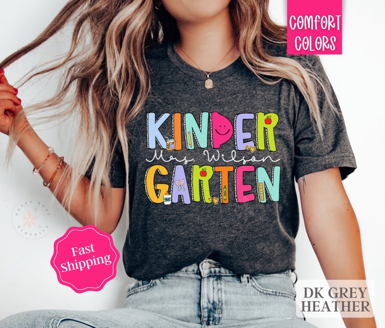 May include: A dark grey heather t-shirt with the text "KINDERGARTEN" in colorful letters with illustrations of school supplies. The text "Mrs. Wilson" is written in black below the word "KINDERGARTEN".