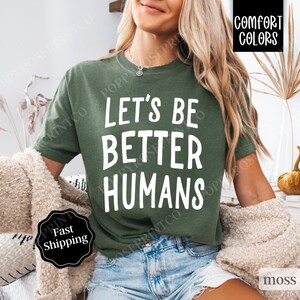 May include: A woman wearing a green t-shirt with the text "Let's Be Better Humans" in white. The shirt is a comfortable fit and has a relaxed style.