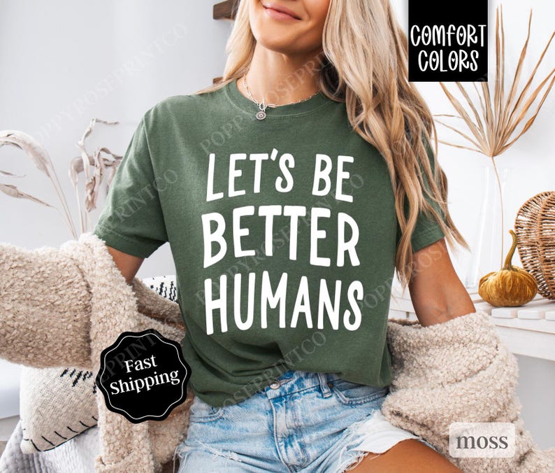 May include: A woman wearing a green t-shirt with the text "Let's Be Better Humans" in white. The shirt is a comfortable fit and has a relaxed style.