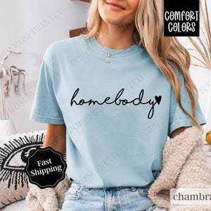 May include: A light blue t-shirt with the text "homebody" in black script with a small black heart to the right of the word. The shirt is made of a soft, comfortable fabric.