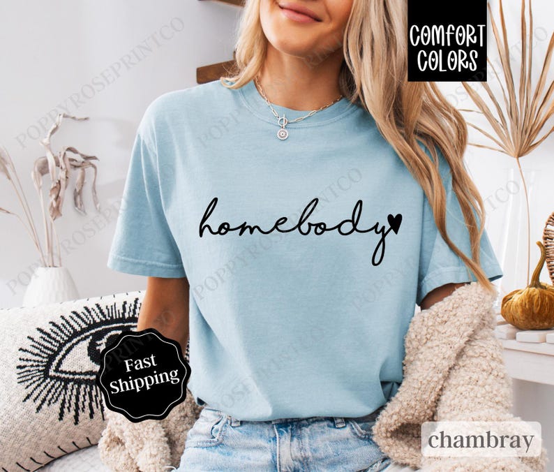 May include: A light blue t-shirt with the text "homebody" in black script with a small black heart to the right of the word. The shirt is made of a soft, comfortable fabric.