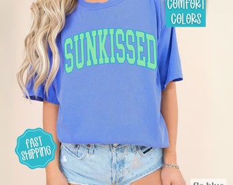 May include: A blue t-shirt with the word "SUNKISSED" in green letters. The shirt is made of a soft, comfortable fabric and has a relaxed fit.