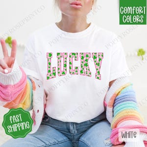 May include: White t-shirt with the word "LUCKY" printed in pink with a green shamrock pattern. The word is in a bold, blocky font.