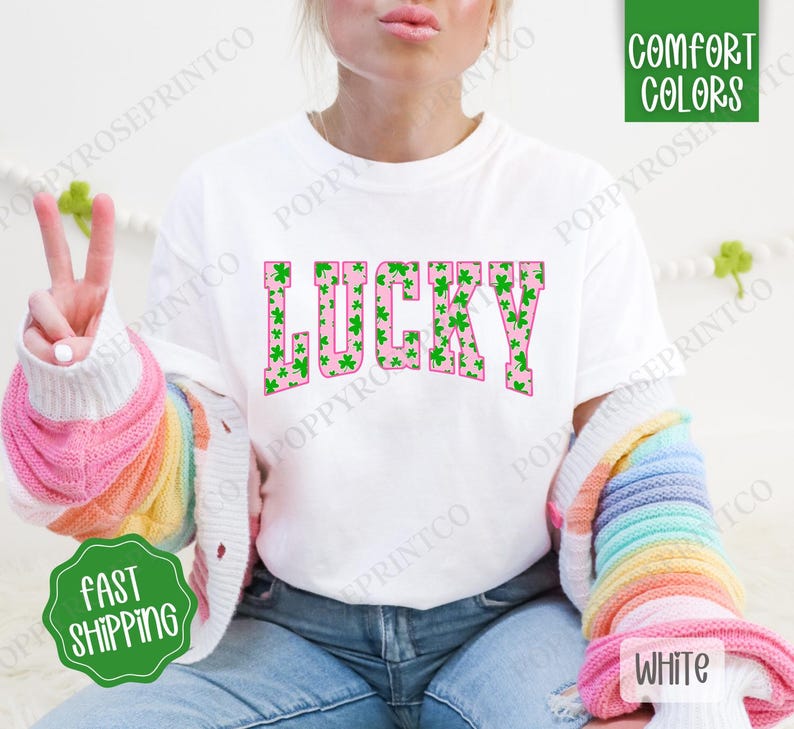 May include: White t-shirt with the word "LUCKY" printed in pink with a green shamrock pattern. The word is in a bold, blocky font.