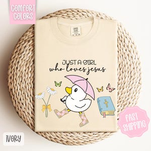 Puede incluir: Camiseta de color marfil con un pato de dibujos animados que lleva botas de lluvia y sostiene un paraguas. El texto "Just a girl who loves Jesus" est&aacute; impreso en la camiseta. Hay una Biblia y flores en el fondo.