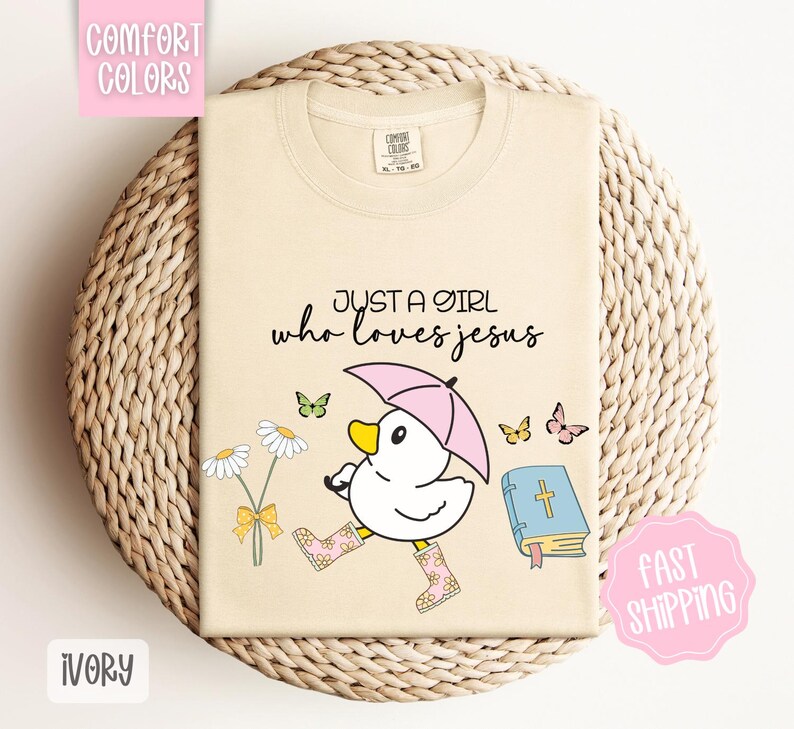 Puede incluir: Camiseta de color marfil con un pato de dibujos animados que lleva botas de lluvia y sostiene un paraguas. El texto "Just a girl who loves Jesus" est&aacute; impreso en la camiseta. Hay una Biblia y flores en el fondo.