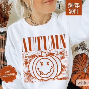 May include: White sweatshirt with the word "AUTUMN" in orange text and a smiling pumpkin graphic with a red outline. The pumpkin has a red tongue sticking out and two red X's for eyes.