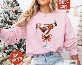 May include: Light pink sweatshirt featuring a graphic of Santa Claus relaxing in a martini glass with a red bow. The sweatshirt has long sleeves and is labeled "SUPER SOFT" and "It pink". The words "FAST SHIPPING" are also visible.