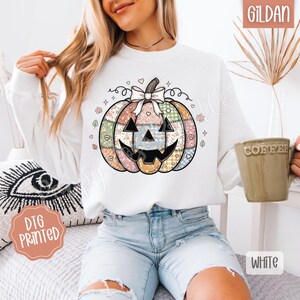 May include: White Gildan sweatshirt featuring a colorful patchwork pumpkin design with a bow and a smiling face. The sweatshirt has the text "DTG Printed" on a peach-colored sticker. The mug says "Coffee".