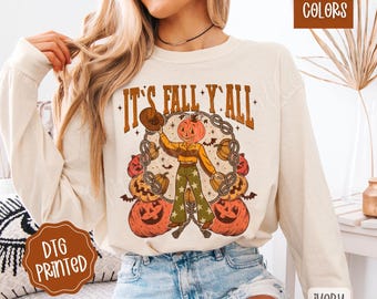 May include: Cream-colored long-sleeve shirt with the text "IT'S FALL Y'ALL" and a graphic of a pumpkin-headed cowboy surrounded by pumpkins. The shirt is printed with a DTG process and is labeled "Comfort Colors" and "Ivory".