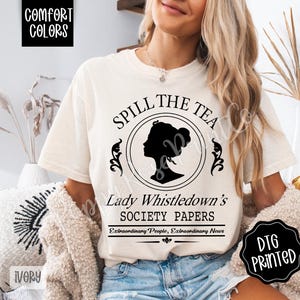 Pu&ograve; includere: T-shirt color avorio con la scritta "Spill The Tea" e "Lady Whistledown's Society Papers" in nero. Il design include una silhouette di profilo femminile all'interno di una cornice circolare. Stampa DTG.