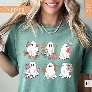 May include: A light green t-shirt with a graphic of six white ghosts holding flowers and butterflies. The ghosts are surrounded by colorful flowers. The shirt has a crew neck and short sleeves.