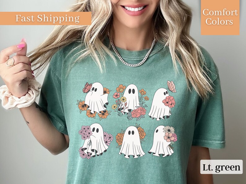 May include: A light green t-shirt with a graphic of six white ghosts holding flowers and butterflies. The ghosts are surrounded by colorful flowers. The shirt has a crew neck and short sleeves.