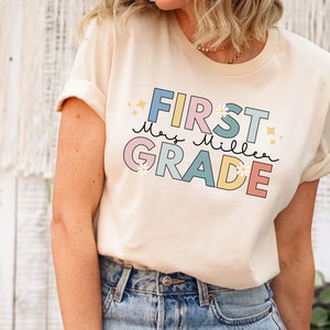 Op de afbeelding: Een licht beige T-shirt met een kleurrijke afbeelding waarop "First Grade" staat, met de naam van de lerares "Mrs. Miller" in cursief. De afbeelding heeft een pastelkleurenpalet met roze, blauw, geel en groen. Er zijn kleine witte bloemen en sterren rond de tekst.