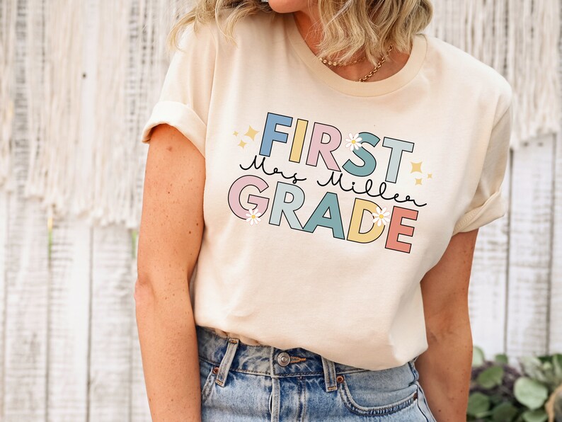 Op de afbeelding: Een licht beige T-shirt met een kleurrijke afbeelding waarop "First Grade" staat, met de naam van de lerares "Mrs. Miller" in cursief. De afbeelding heeft een pastelkleurenpalet met roze, blauw, geel en groen. Er zijn kleine witte bloemen en sterren rond de tekst.