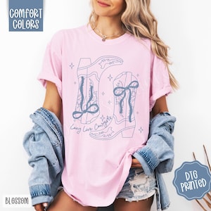 May include: A light pink t-shirt with a blue graphic of cowboy boots and the text "Long Live Cowgirls". The shirt has rolled-up sleeves and is paired with a denim jacket. The words "Comfort Colors" and "Blossom" are visible.