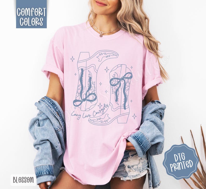May include: A light pink t-shirt with a blue graphic of cowboy boots and the text "Long Live Cowgirls". The shirt has rolled-up sleeves and is paired with a denim jacket. The words "Comfort Colors" and "Blossom" are visible.