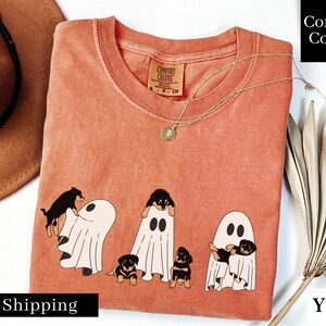 May include: A peach-colored t-shirt with a graphic of four black and brown puppies dressed as ghosts. The text "Comfort Colors" is printed on the tag.