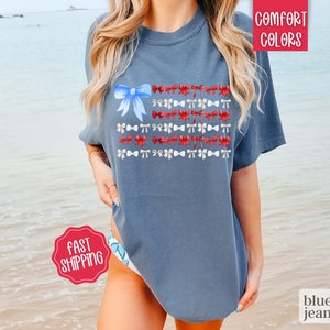 May include: Blue jean colored t-shirt with a watercolor design of a flag made of bows. The flag design features a large blue bow in the upper left corner and rows of red and white bows. The words "Comfort Colors" are in the upper right corner.